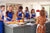 Group of people wearing blue aprons standing around a chef learning to cook.